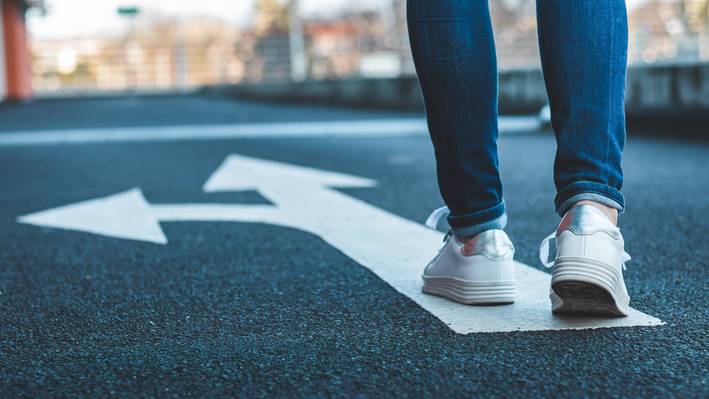Für Profis und Quereinsteigende Zwei Füsse stehen auf der Strasse auf einem Pfeil der geradeaus zeigt und eine Abzweigung nach links hat.
