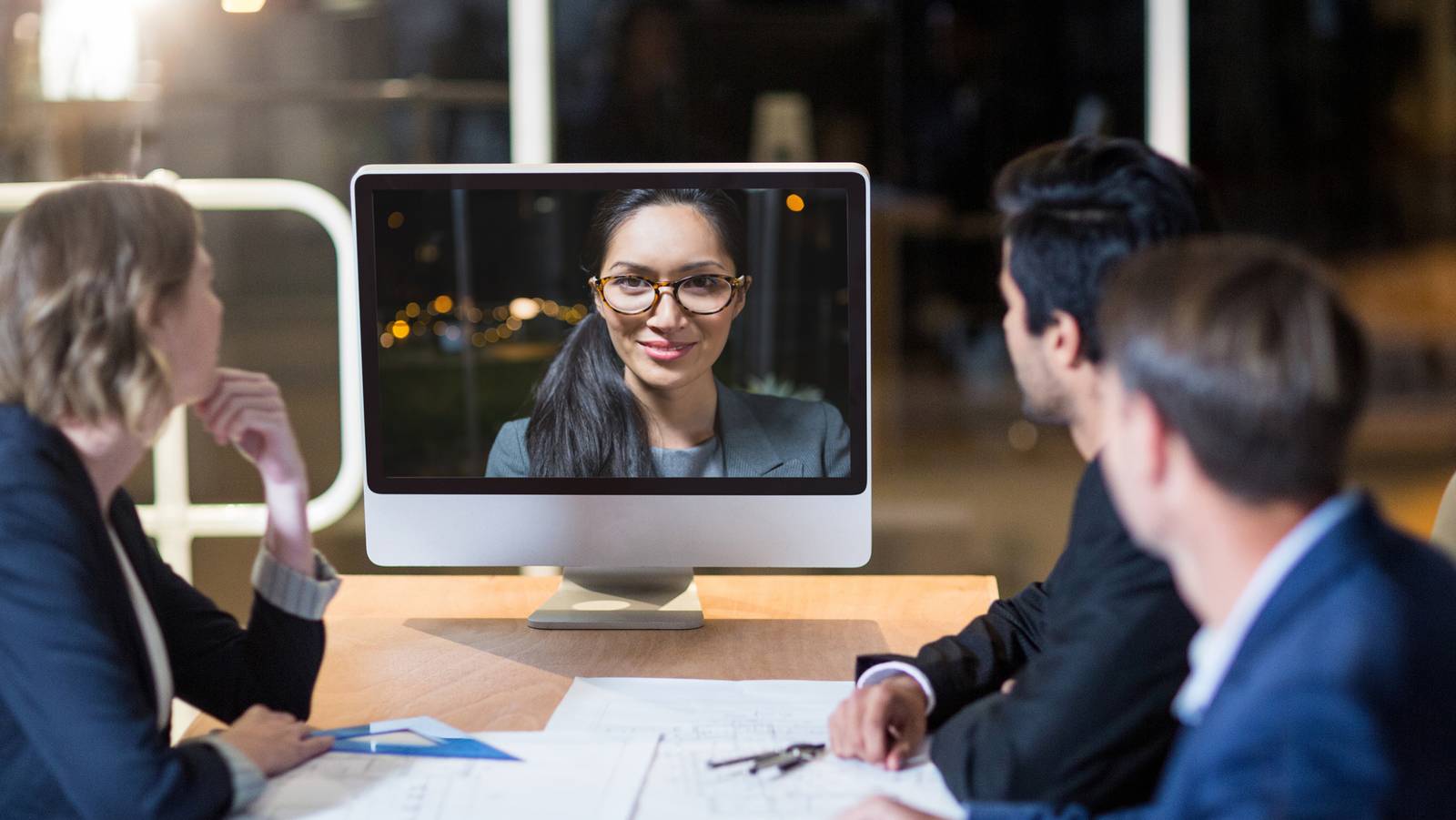 Bachelor Thesis Personalarbeit Personen an einem Konferenztisch, die auf einen Monitor blicken in dem eine Frau virtuell an der Sitzung teilnimmt